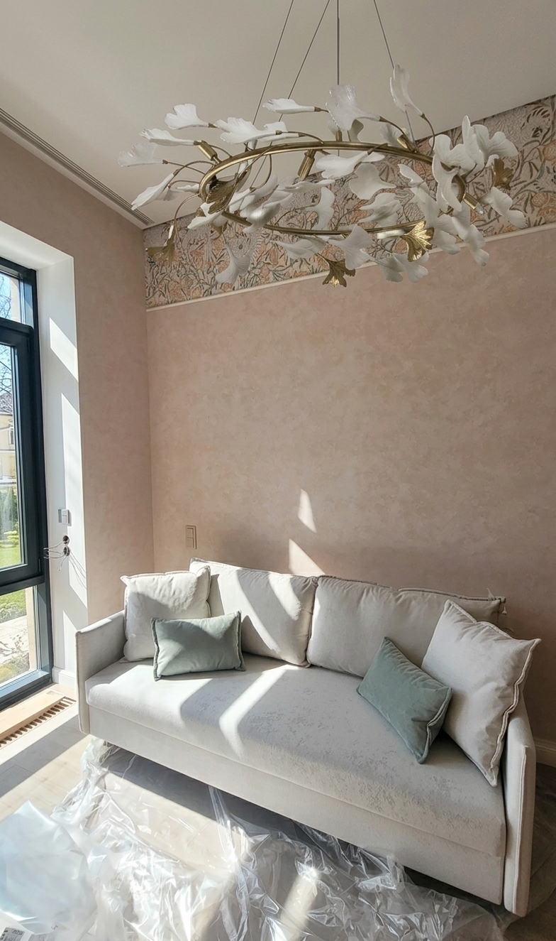 A crisp midday interior photograph captured with a 35mm lens, where bright, natural spring daylight streams through the tall window on the left, casting sharp geometric sunbeams and soft ambient bounce across the scene. Maintain the exact composition, geometry, and placement of the plush light-beige sofa, muted accent pillows, textured blush-toned plaster wall, floral frieze, and the sculptural white-and-brass ginkgo leaf chandelier. Render authentic physical textures, including the tactile fabric weave of the upholstery, subtle crinkling and sheen on the plastic floor covering, metallic glints on the chandelier frame, and natural surface irregularities in the wall finish, balanced with gentle photographic grain, realistic depth of field, and faint lens flare.