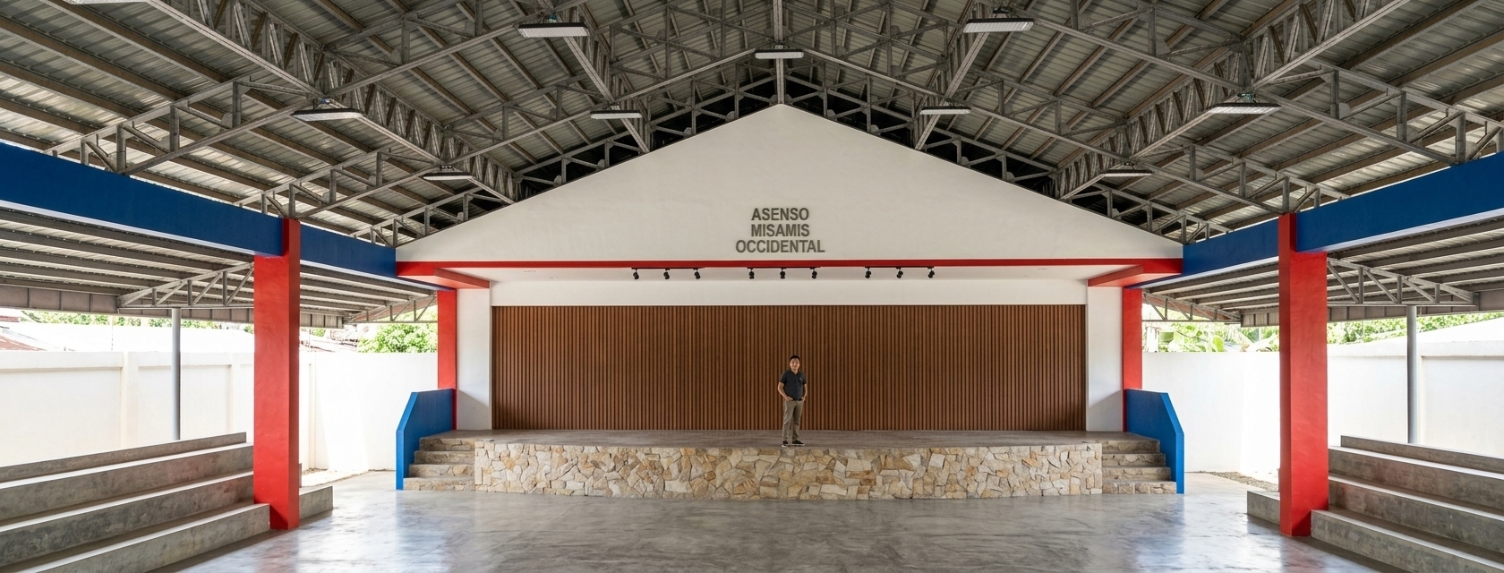 A wide-angle architectural photograph capturing this exact covered gymnasium scene during a bright summer midday. Crisp, overhead tropical sunlight filters beneath the roof edges, casting harsh, realistic shadows across the smooth concrete floor and bleachers while filling the interior with warm ambient bounce. The steel roof trusses display authentic matte metallic textures and structural welds, complementing the corrugated metal decking above. The stage features a warm, vertically paneled natural wood backdrop, textured concrete risers, and matte red-painted columns showing subtle surface imperfections. The figure on stage is rendered as an authentic person with realistic fabric draping, under a balanced exposure with subtle chromatic aberration, sharp focus, and zero distortion of the original composition.