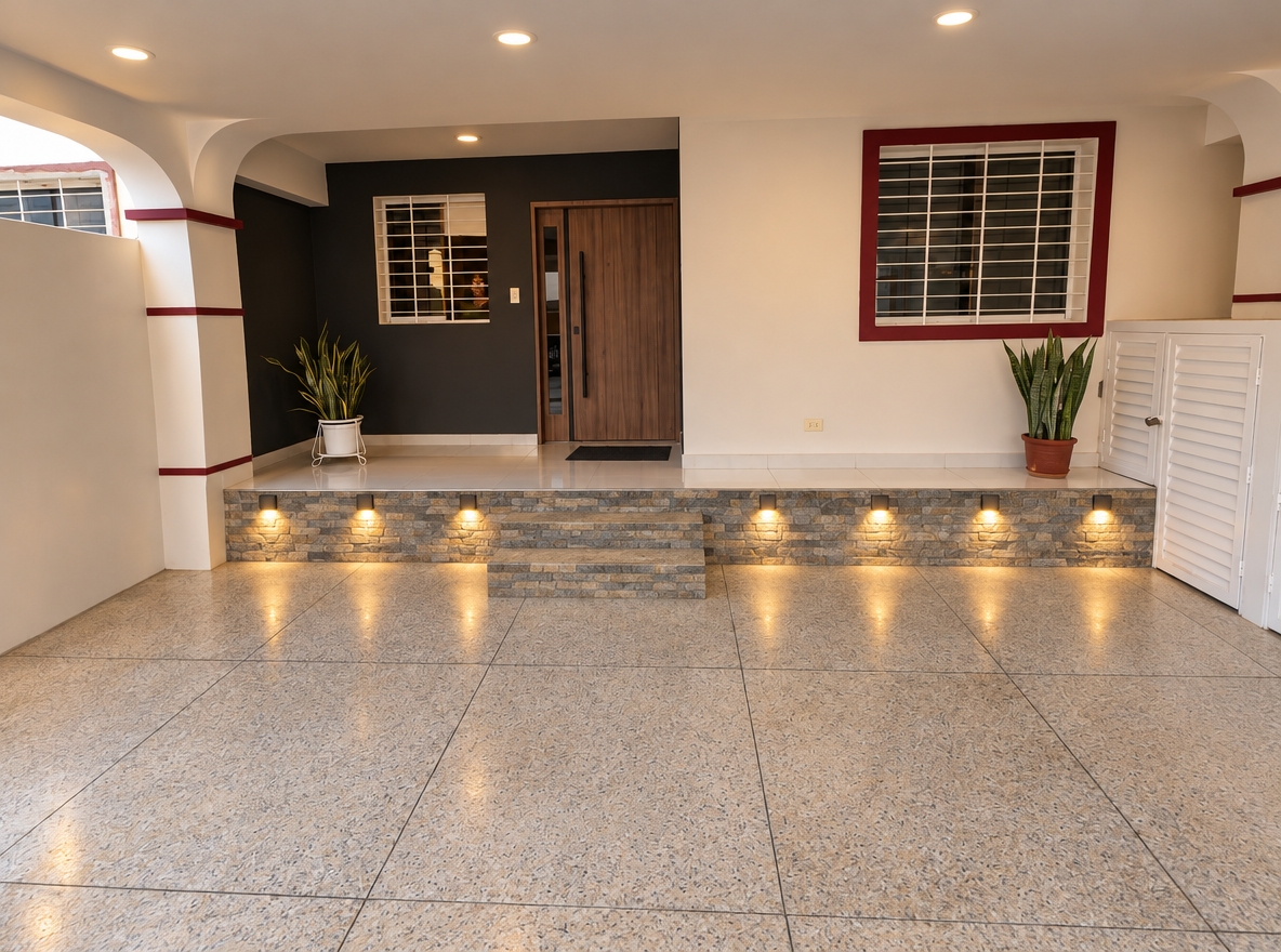 An incredibly lifelike, richly detailed, photorealistic wide-angle shot of a contemporary residential entrance set in a private, clean-lined urban courtyard. The scene is illuminated by soft, inviting warm-toned LEDs recessed into the stone base, creating a serene and sophisticated atmosphere. Captured with a 24mm wide-angle lens, emphasizing the crisp textures of the natural stacked-stone masonry and the sleek contrast between the matte charcoal and crisp white wall finishes.
The architectural design features a raised porch elevation clad in textured "espacato" natural stone, which now seamlessly spans the entire base, replacing the original lighting fixture with a continuous, uniform masonry surface. Seven square, recessed warm-light luminaires are integrated horizontally along the stone face, casting precise, downward-directed beams that highlight the earthy variations of the rock face. A modern, vertical-grain wood door serves as the focal point, flanked by white-walled sections and guarded by window security grilles finished in a striking deep red. The composition is grounded by high-quality granite-flecked floor tiles that extend across the driveway, while two vibrant snake plants add a touch of organic life, reinforcing the home’s sense of contemporary elegance and inviting human scale.