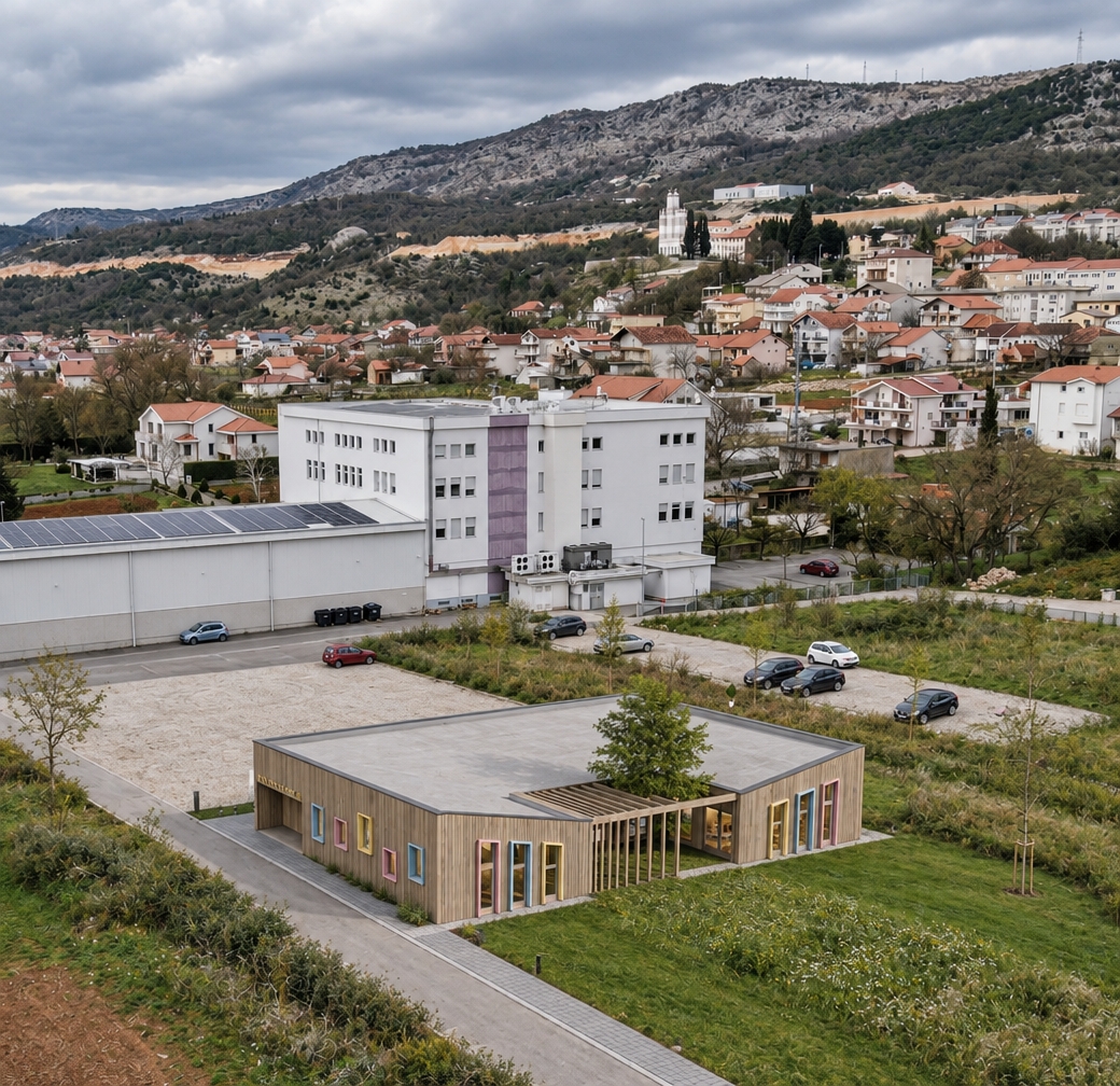 A high-resolution aerial photograph capturing the exact scene under soft, overcast daylight, highlighting the natural grain and tactile matte finish of the kindergarten’s Scandinavian vertical timber cladding and pastel window trims. The surrounding grounds, asphalt driveway, and gravel parking areas display authentic photorealistic textures with subtle dust, wear, tire marks, and natural ground variations. The grassy lawn in front features organic blade variations and wildflowers, while the background hillside, village rooftops, and rugged mountain reveal crisp depth, natural atmospheric haze, balanced exposure, and true-to-life colors.